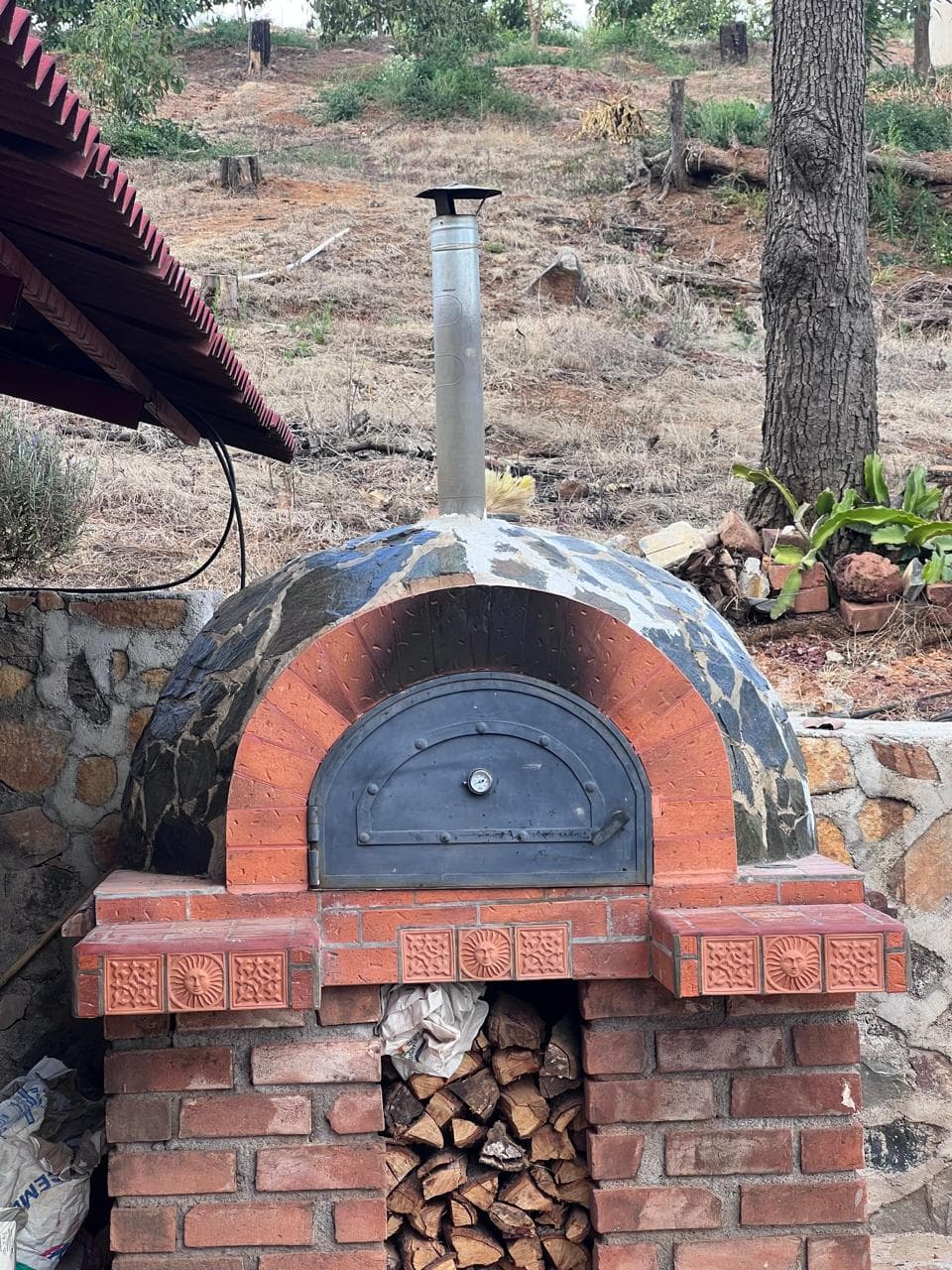 Vista frontal de un horno artesanal de leña construido con ladrillo, piedra, puerta metálica y espacio inferior para almacenar madera.
